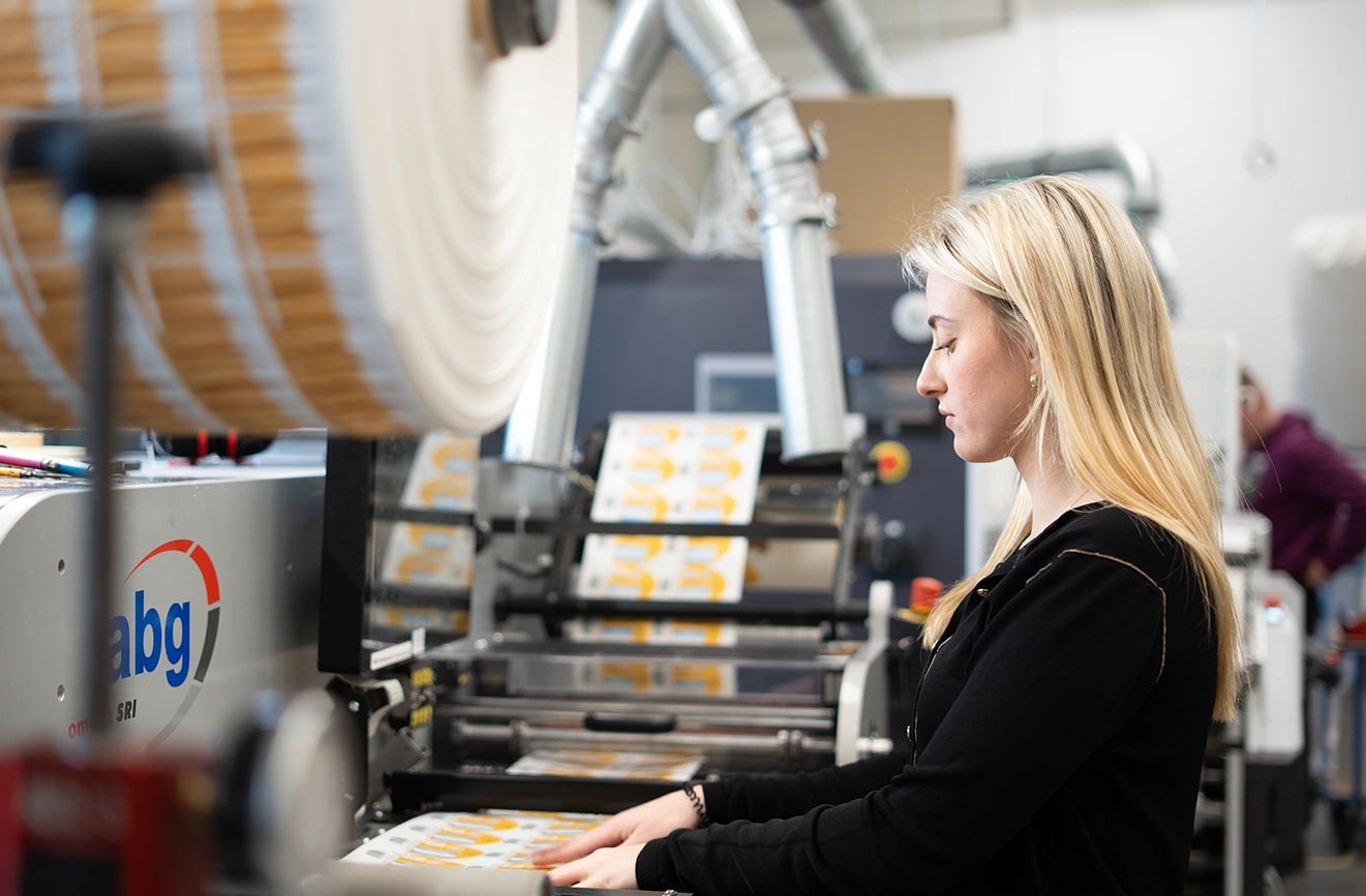 Eine Mitarbeiterin bedient eine industrielle Etikettendruckmaschine und führt bedruckte Etikettenbögen. Im Vordergrund sind große Materialrollen zu sehen, während die Produktionsanlage und weitere Maschinen im Hintergrund unscharf dargestellt sind.