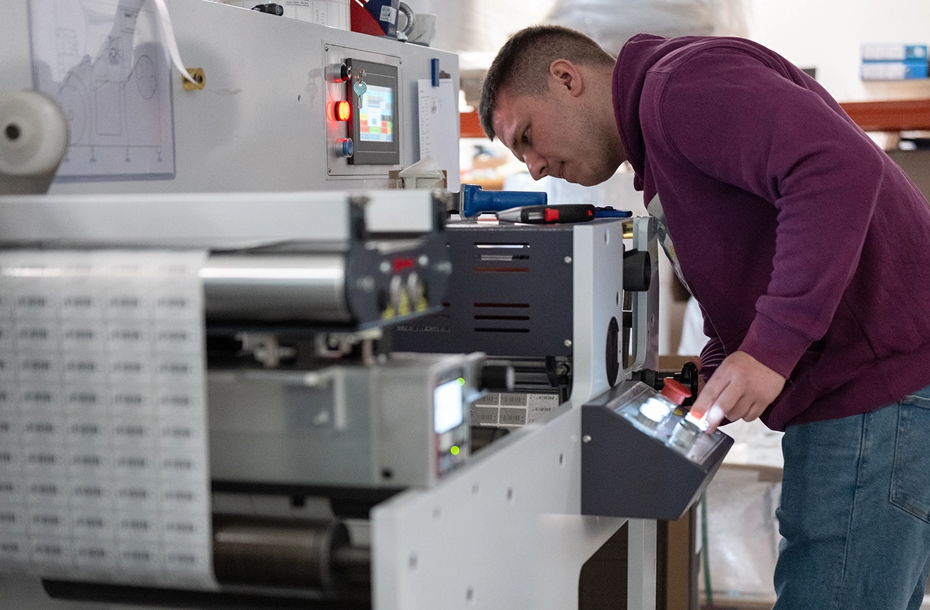 Ein Mitarbeiter bedient eine industrielle Etikettendruckmaschine und überprüft die Einstellungen am Bedienfeld. Im Vordergrund sind bedruckte Etiketten auf einer Materialbahn sowie Teile der Produktionsanlage zu sehen.