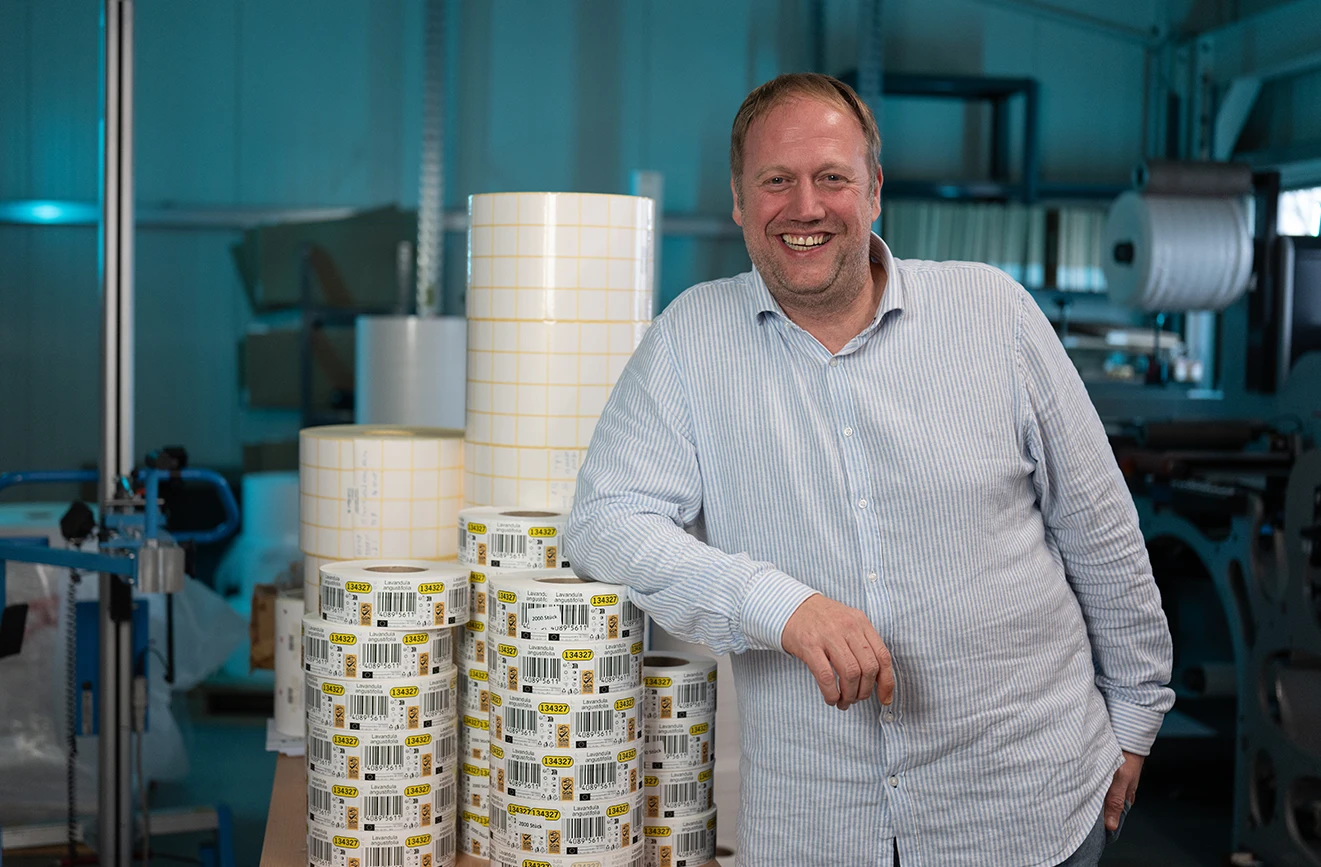Ein lächelnder Mann steht in einer Produktionshalle neben mehreren Rollen mit bedruckten Etiketten. Im Hintergrund sind Maschinen, große Materialrollen und weitere Produktionsanlagen einer Etikettendruckerei zu sehen.